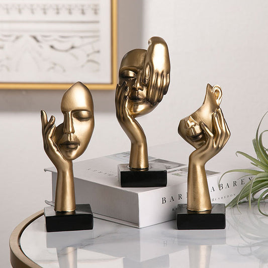 Three gold face sculptures on black bases on a reflective surface with a blurred background