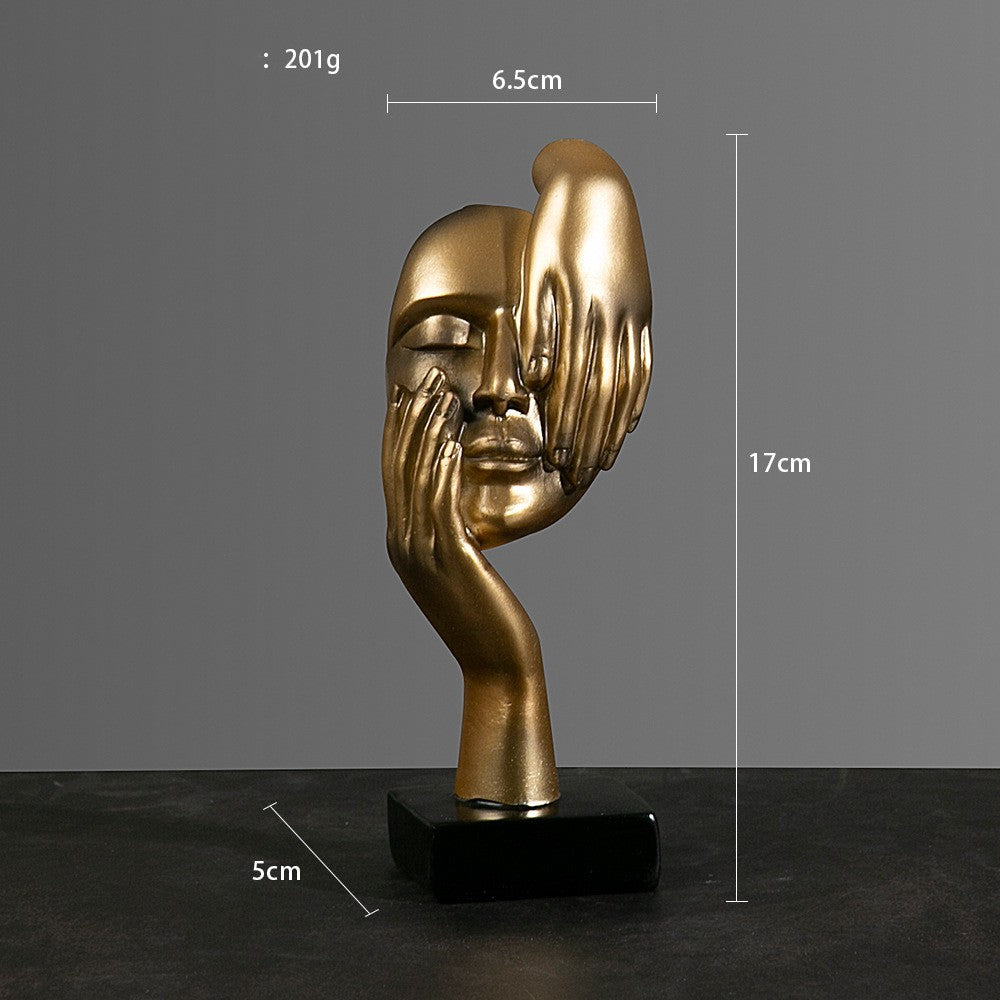 Gold sculpture of a face with hands on a black base against a gray background