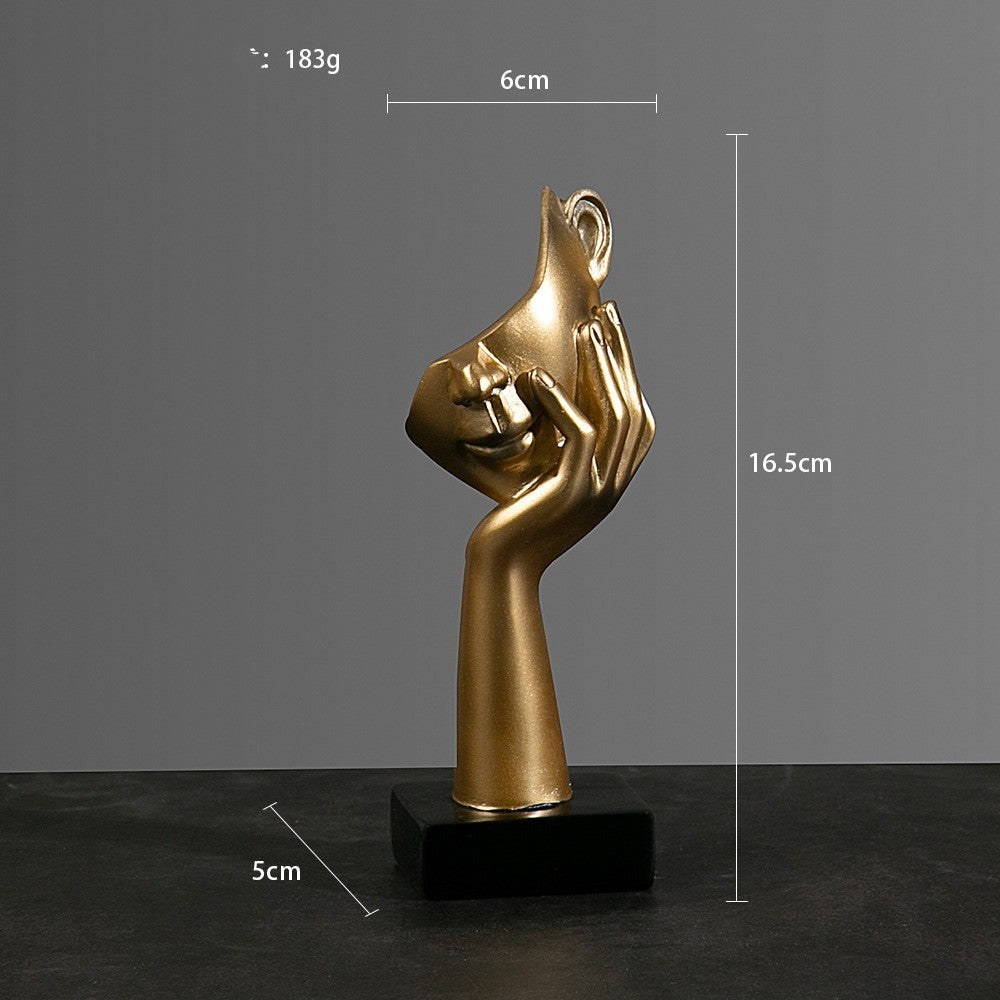 Gold sculpture of a hand holding a face on a black base with measurements against a gray background